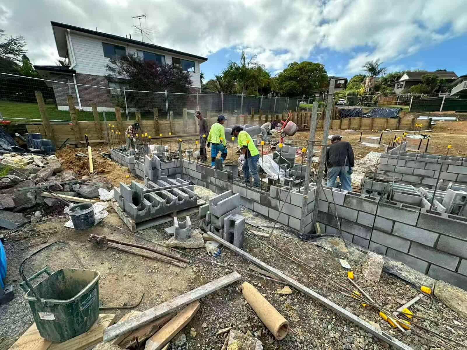 Workers laying a reinforced concrete block wall on a subdivision site, with starter bars visible in the cores